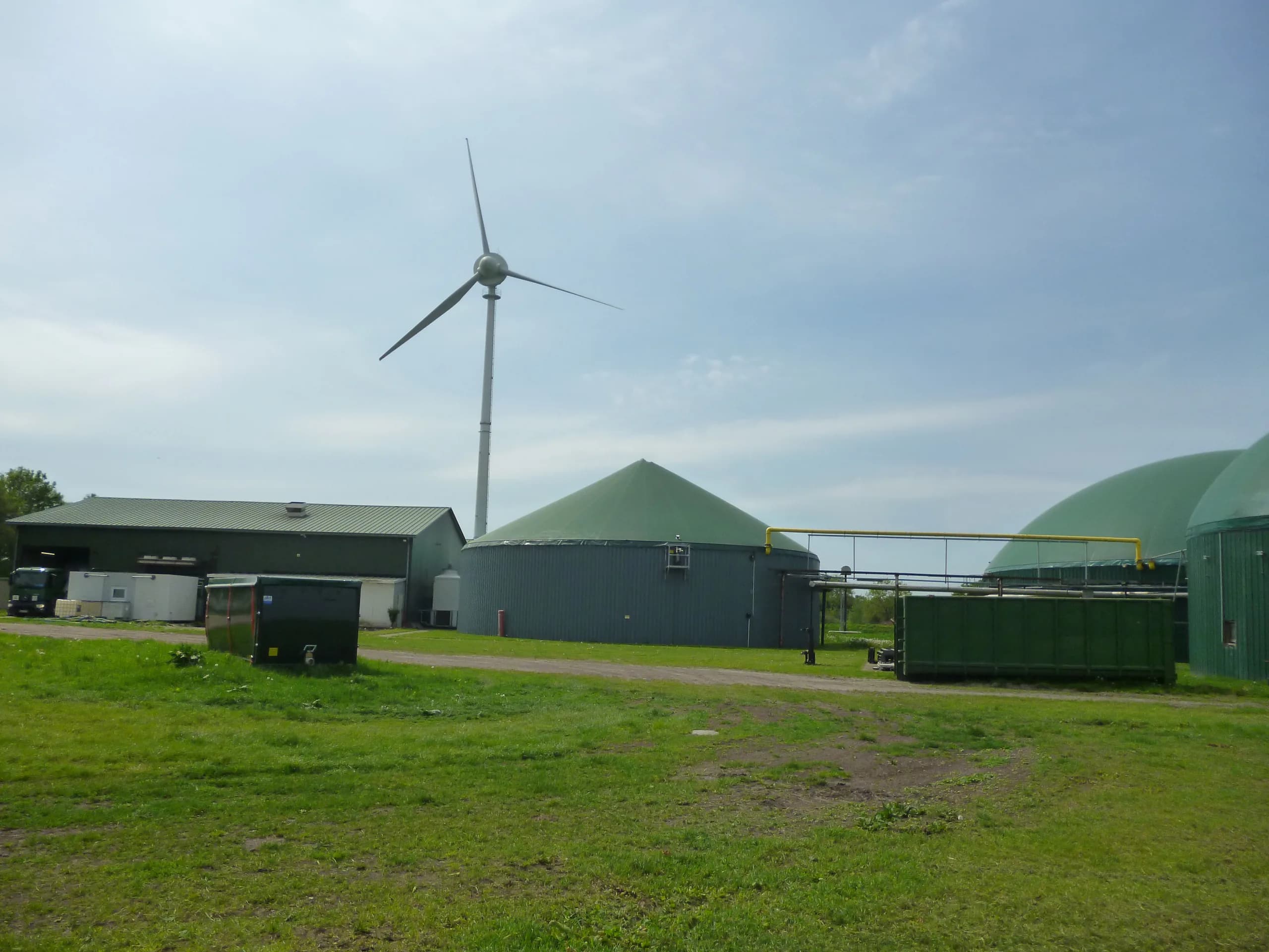 Biogasanlage mit grün überdachten Behältern, Gasleitung und Windrad im Hintergrund